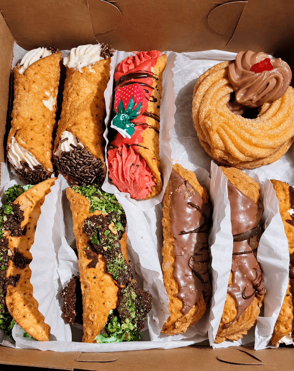 Assortment of Italian bakery pastries including cannoli and chocolate-glazed pastries in a bakery box